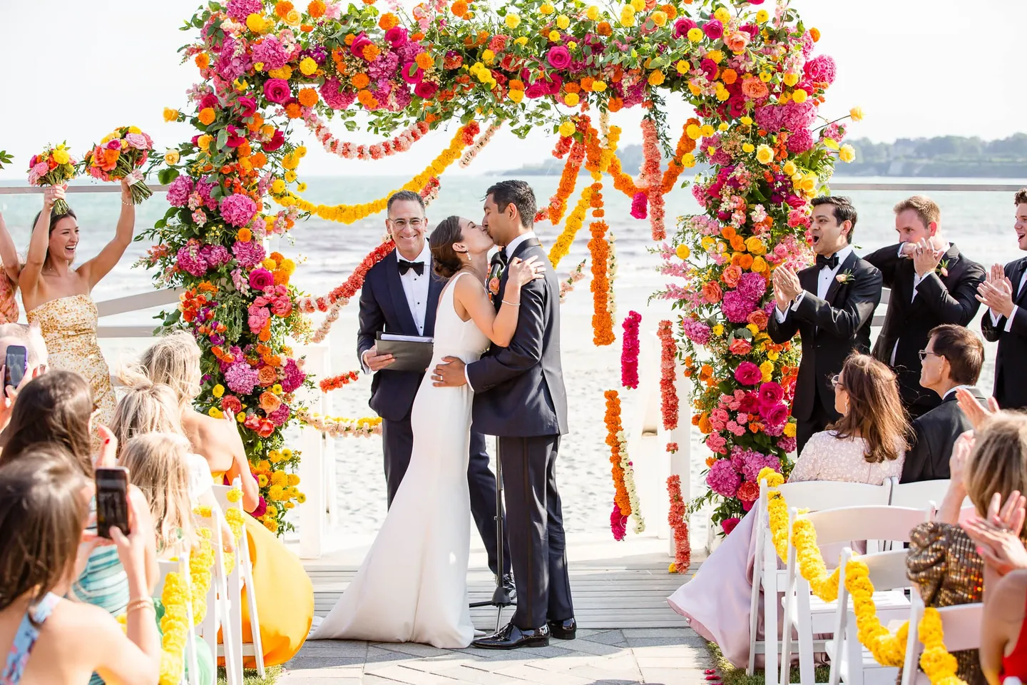 Zoey and Akshay kiss under the marigold arch as the wedding party cheers