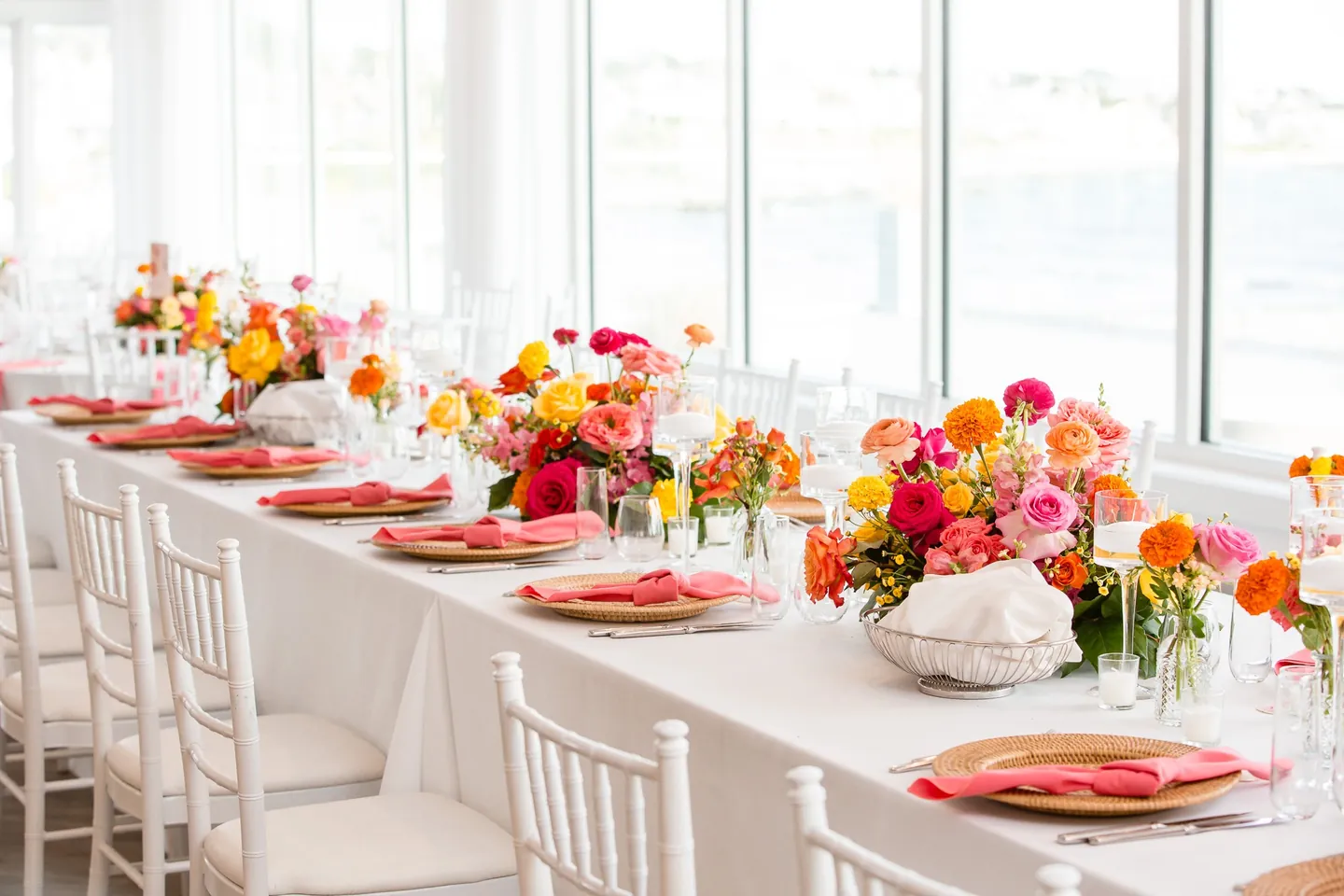 Long reception tables with coral napkins, gold chargers and orange centerpieces by tall windows