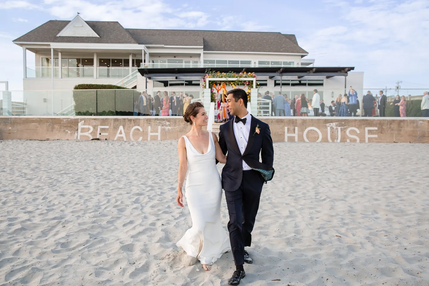 Zoey and Akshay walk across the sand in front of the Beach House sign wall