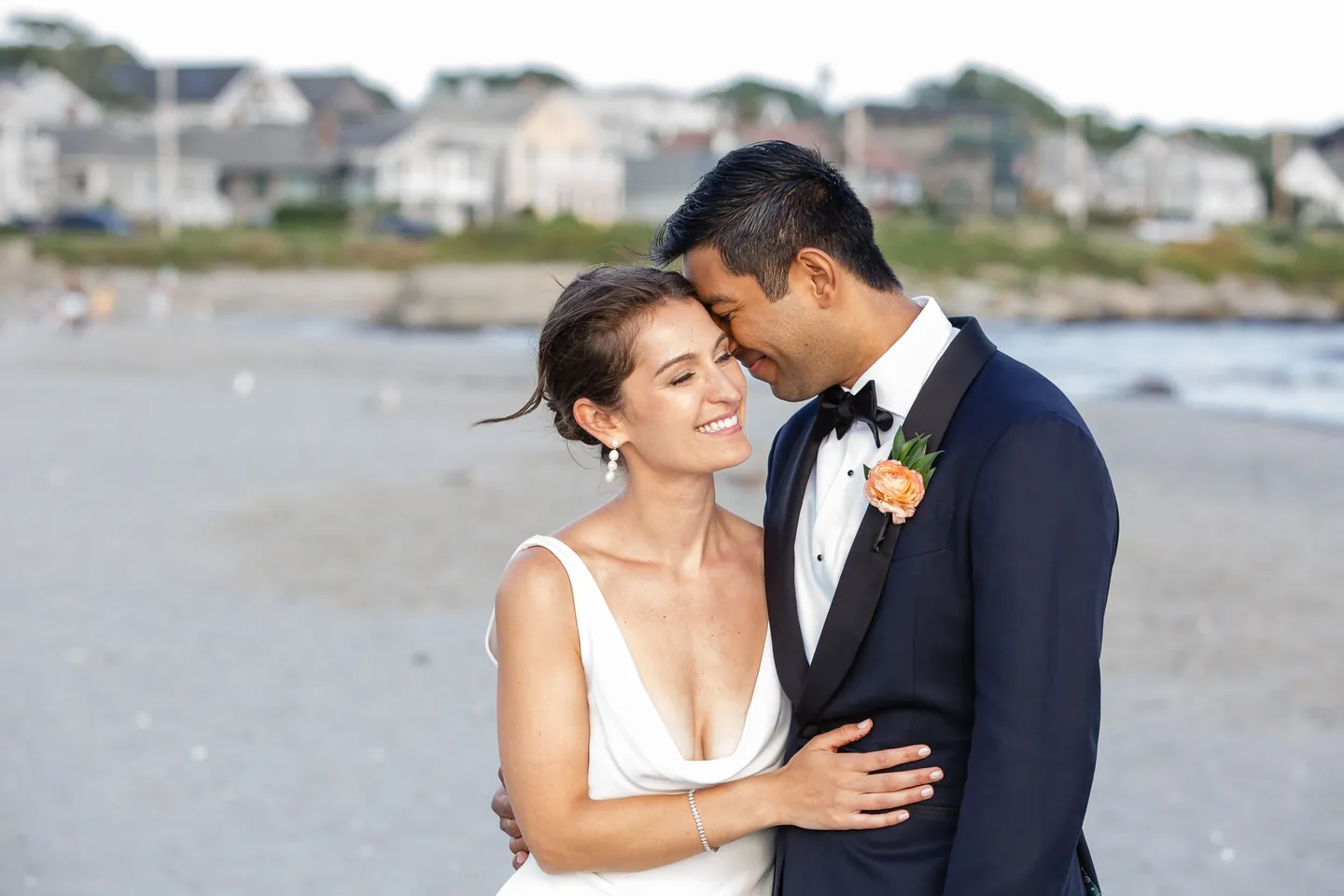 Zoey smiles with her head against Akshay's on the beach, shingled houses soft behind