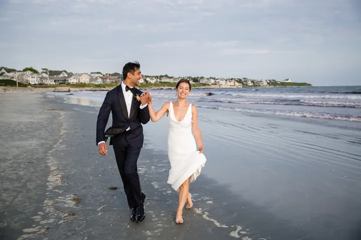 Zoey and Akshay run barefoot along wet sand at the waterline, her hem gathered in hand