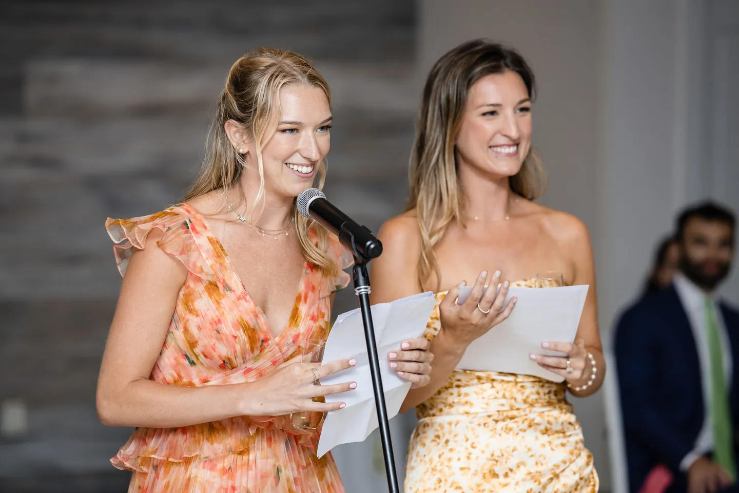 Two bridesmaids in metallic and floral dresses read a toast from paper at the microphone